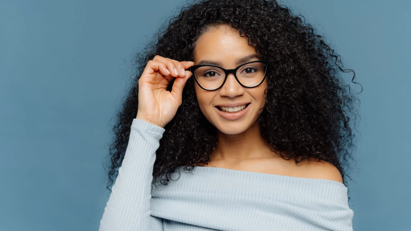 A smiling woman with curly hair adjusting her black-framed glasses